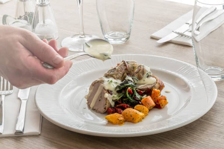 Hand pouring white sauce over pork and vegetables on plate